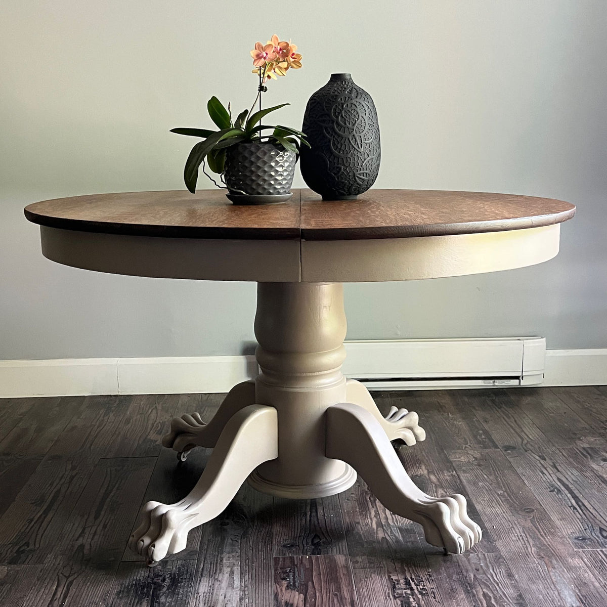 A round pedestal dining table with a medium brown wood top and a distressed beige/cream painted base with claw legs, shown on a dark wood floor against a light gray wall. Decorative items (a potted orchid and a textured vase) are placed on top.