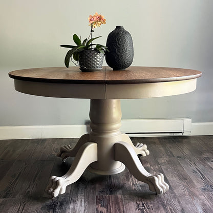 A round pedestal dining table with a medium brown wood top and a distressed beige/cream painted base with claw legs, shown on a dark wood floor against a light gray wall. Decorative items (a potted orchid and a textured vase) are placed on top.