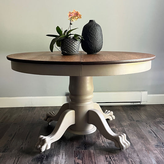 A round pedestal dining table with a medium brown wood top and a distressed beige/cream painted base with claw legs, shown on a dark wood floor against a light gray wall. Decorative items (a potted orchid and a textured vase) are placed on top.